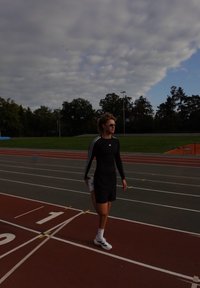 Atleta vestido com uma camisola de mangas compridas preta com riscas brancas, calções pretos e sapatos brancos, a alongar-se numa pista de corrida sob um céu nublado.