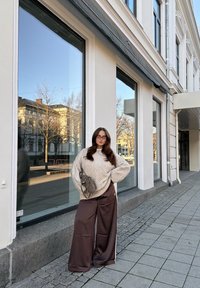 Chunky beige strikket sweater, brune vidde ben bukser med sidelommer, bærer en lille grå taske; står på en brolagt fortov nær store vinduer.
