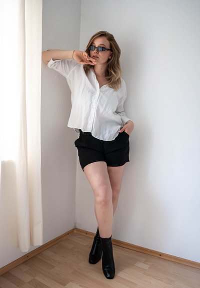 Camisa blanca de lino con botones, pantalones cortos negros y botines negros. El modelo se apoya contra una pared con un fondo neutro y luz natural.