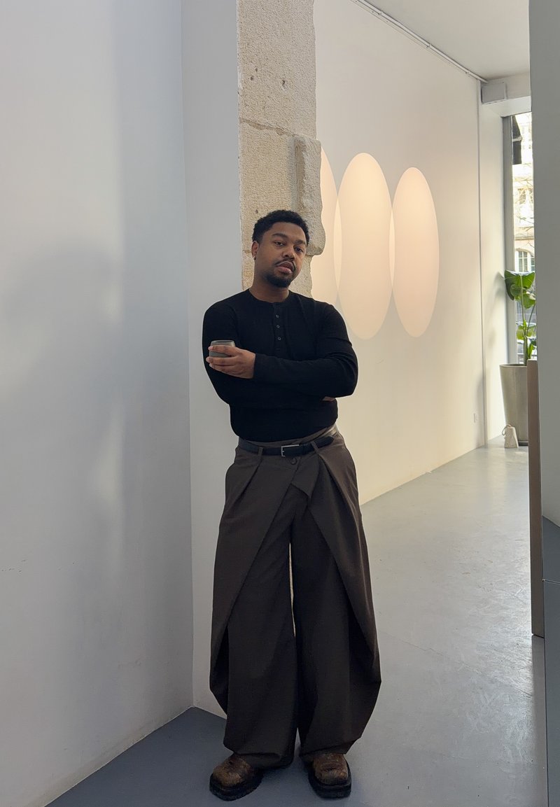 Mann mit verschränkten Armen, der ein Telefon hält, trägt ein schwarzes Langarmshirt und breite braune Hosen, steht in einem minimalistischen Galerieraum mit Lichtkunst.