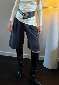 Personne portant un haut blanc à manches longues, une large ceinture noire avec une boucle ronde, un short noir ample arrivant aux genoux, et des bottines noires brillantes à lacets avec un talon moyen.