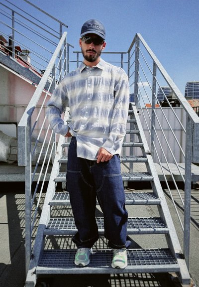 Camisa de botones con rayas azul claro y blancas; jeans desgastados de mezclilla oscuros y holgados; zapatillas gris con acentos verdes; gorra de mezclilla; gafas de sol; de pie en unas escaleras.