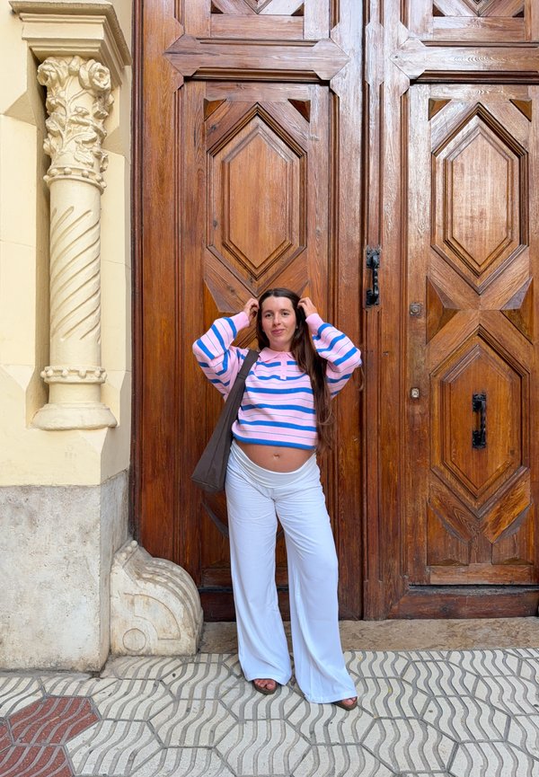 Joven mujer con cabello largo, con una camiseta a rayas rosa y azul y pantalones blancos, está de pie frente a grandes puertas de madera ornamentadas.