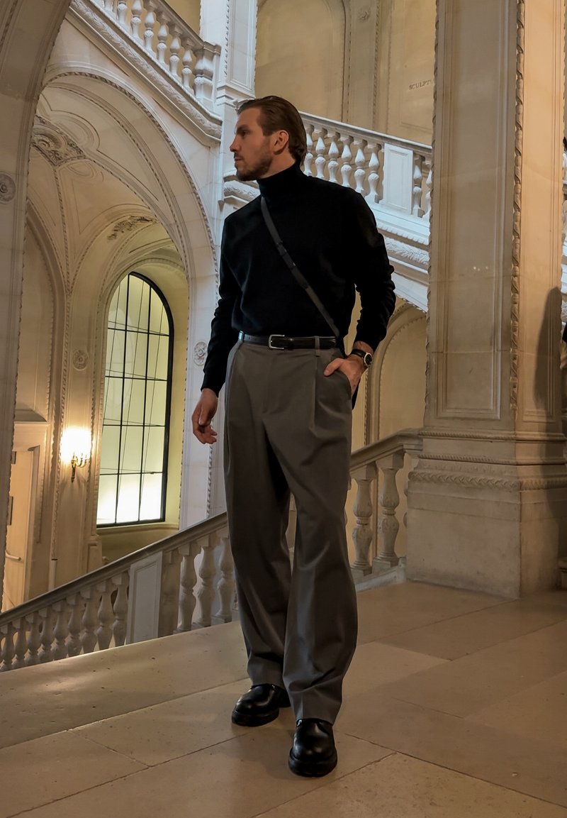 Maglione a collo alto nero, pantaloni grigi a pieghe e stivali neri. L'outfit presenta un design minimalista con una borsa a tracolla in pelle e un orologio da polso.