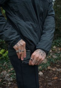 Casaco preto texturizado com bolsos, usado por uma pessoa a inserir um smartphone no bolso. Mãos adornadas com anéis e tatuagens.
