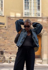 Τζάκετ τζην με χαλαρή εφαρμογή σε μπλε, φορεμένο πάνω από μαύρη μπλούζα και χαλαρούς μαύρους παντελόνες. Γυαλιά ήλιου και καφέ τσάντα. Τοίχος από πέτρα στο φόντο.