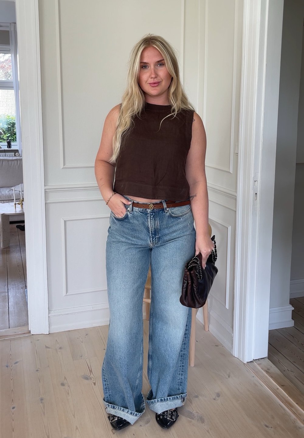 Hnědý top bez rukávů, s vysokým límcem, spárovaný se světle modrými rozšířenými džíny. Drží tmavě hnědou kabelku. Černé špičaté boty dokončují outfit.