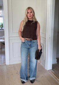 Hnědý top bez rukávů, s vysokým límcem, spárovaný se světle modrými rozšířenými džíny. Drží tmavě hnědou kabelku. Černé špičaté boty dokončují outfit.