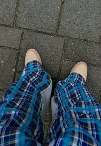 Mocassins bejes com bordas onduladas combinados com calças de padrão quadriculado azul e preto com realces verdes e brancos, sobre um pavimento cinza texturizado.