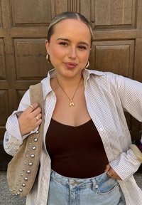 Eine Frau trägt ein gestreiftes, übergroßes weißes und hellbraunes Hemd über einem braunen Tanktop, kombiniert mit hellblauen Denimshorts und einer beigefarbenen Tasche mit silbernen Nieten.