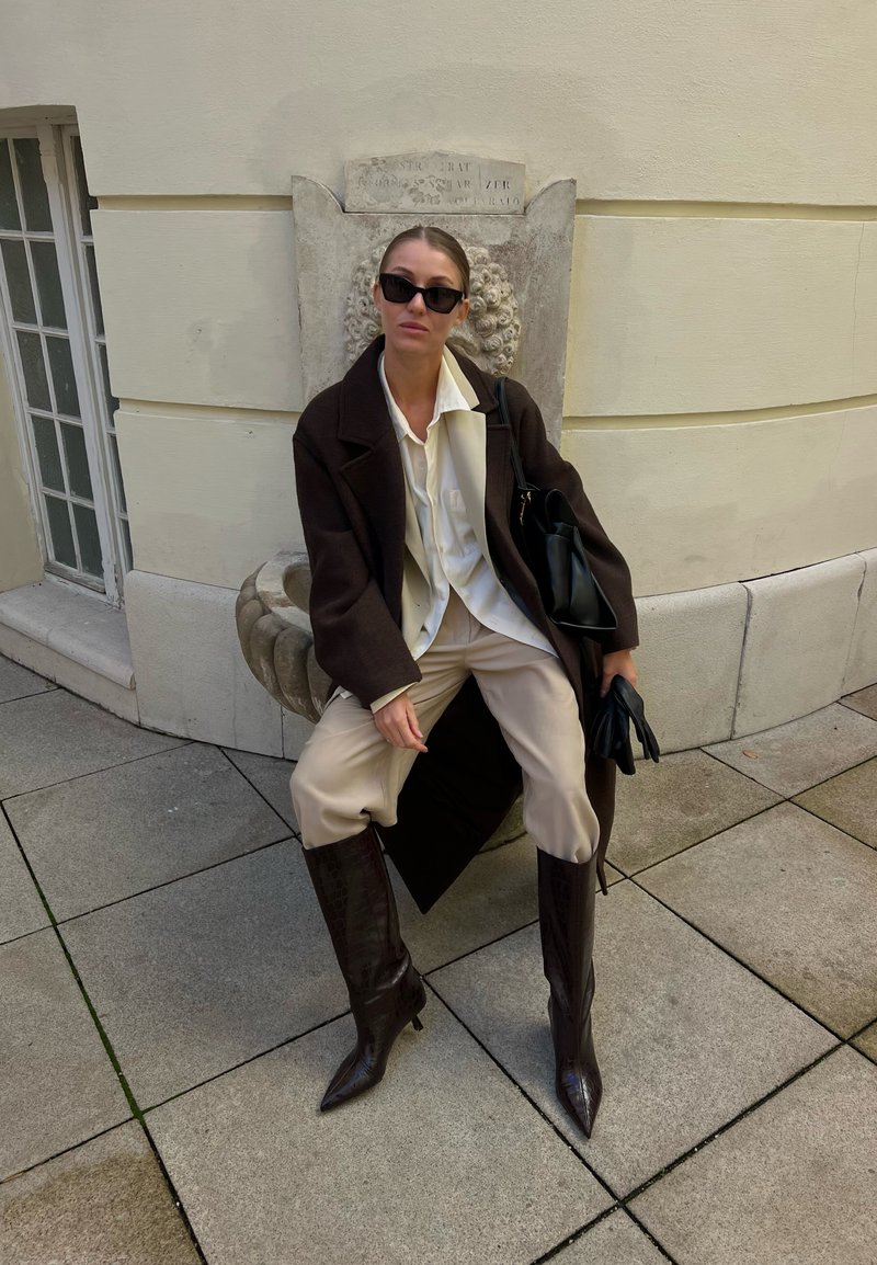 Brown oversized coat, cream shirt, beige trousers, and glossy knee-high brown boots. A black handbag and gloves complete the outfit.