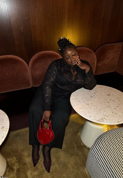 Blusa de encaje negro con cuello alto, pantalones sueltos negros, bolso en forma de corazón rojo y zapatos puntiagudos burdeos frente a un fondo texturizado.