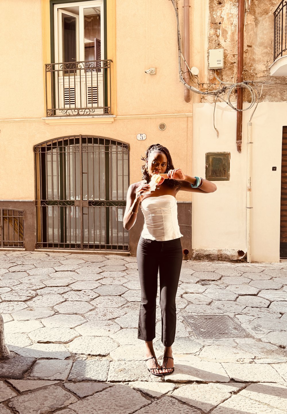 White strapless top, black cropped pants, and flip-flops. Standing on cobblestone, holding an ice cream cone in front of a building.
