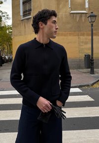 Homme portant un pull noir à fermeture éclair et un pantalon foncé, tenant des gants noirs, debout sur un passage pour piétons en ville près d'un bâtiment jaune et d'un lampadaire.