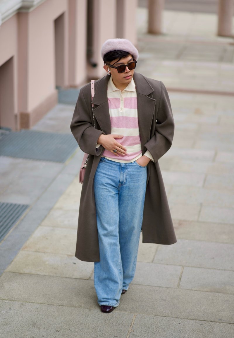 Persona che indossa un cappotto oversized marrone, una camicia a righe rosa e bianca, jeans azzurri e un berretto rosa. Occhiali da sole scuri e auricolari visibili.