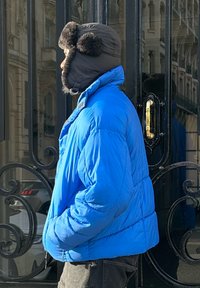 Blå dunjakke med høj krave, der har et glansfuldt stof og en løs pasform. Sort hat foret med pels og pomponer. Mørke bukser, set fra siden.