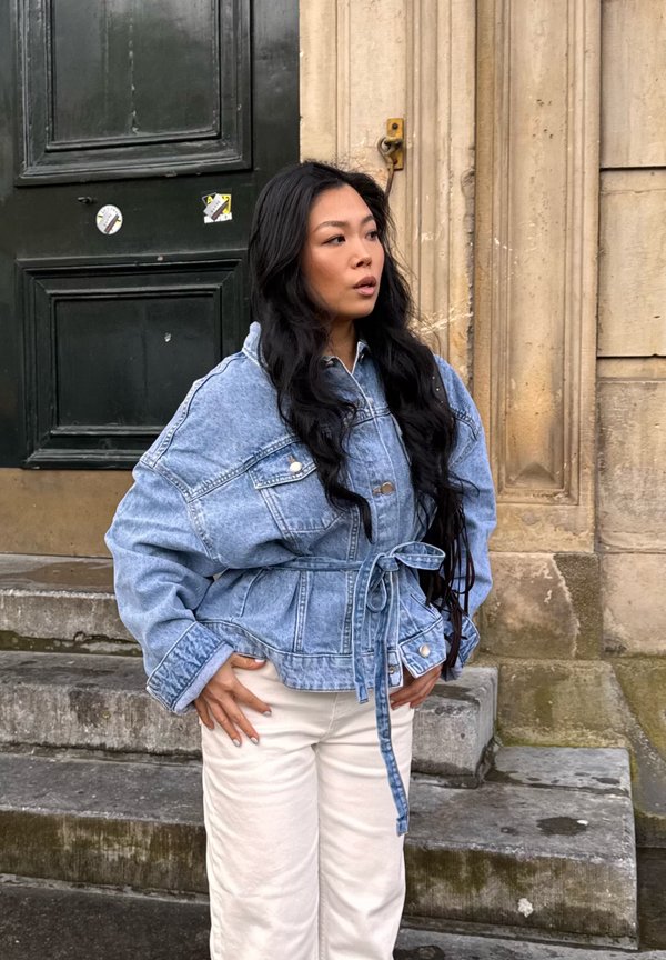 Femme aux longs cheveux noirs portant une veste en jean bleue ceinturée et un pantalon blanc, debout sur des marches en pierre près d'une grande porte sombre et d'un mur en pierre beige.