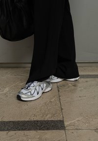 Ezüst és fehér sportcipők hálós felsőrésszel, texturált ezüst díszítéssel és fekete fűzőkkel, fekete nadrággal és fekete táskával párosítva.