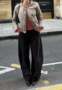 Helle beigen Bomberjacke mit rundem Kragen, kombiniert mit einem rot-weiß gestreiften Hemd und weitem schwarzen Hosen, ergänzt durch schwarze Schuhe.