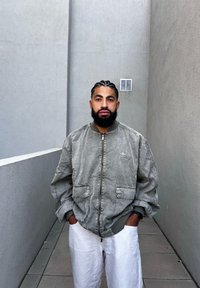 Light grey bomber jacket with ribbed cuffs and zipper pockets, paired with white wide-legged pants, standing in a narrow, minimal space.