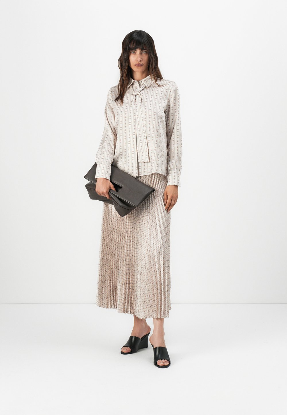 Chemise à manches longues à motif léger beige et marron, associée à une jupe plissée beige. Tenant une pochette grise, portant des mules à talons noirs.