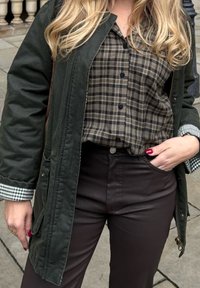 Olivgrüne Zip-up Jacke über einem braun-schwarz karierter Hemd, kombiniert mit dunkelbraunen Hosen. Der Ärmel des Hemdes hat ein grünes Gingham-Muster.