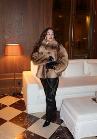 Giacca marrone in pelliccia sintetica con una forma squadrata, pantaloni in pelle nera con dettaglio a catena dorata e stivali appuntiti in pelle nera.