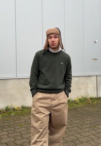 Zöld kötött pulóver, bézs bő nadrág és világosbarna, szőrmével bélelt sapka fülvédővel. Egy aszfaltozott felületen áll, egy szürke fal közelében.