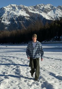 Personne portant une chemise en flanelle à carreaux bleus et blancs, un pantalon vert olive et un bonnet marron clair marchant sur un sol enneigé avec des montagnes en arrière-plan.