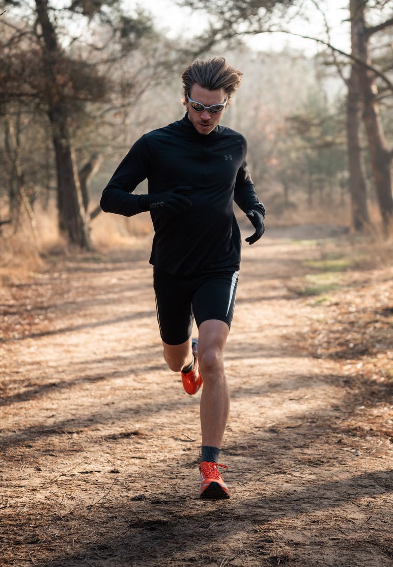Coureur en haut noir à manches longues et short, portant des lunettes de soleil et des gants, sprintant sur un sentier de terre avec des arbres en arrière-plan.
