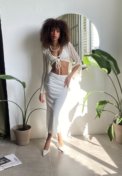 Top corto de encaje blanco con detalle de lazo en el frente, combinado con una falda maxi blanca y tacones blancos puntiagudos. Plantas verdes y un espejo redondo en el fondo.