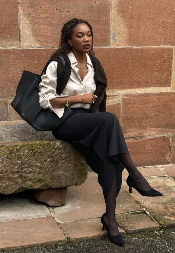 Donna con i capelli intrecciati seduta su una cornice di pietra che indossa una camicetta bianca, una gonna nera, tacchi neri e porta una grande borsa nera.