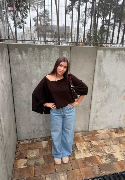 Mujer joven con cabello largo y castaño, de pie con las manos en las caderas, lleva una blusa suelta negra, vaqueros anchos de pierna en azul claro y zapatos beige junto a una pared de hormigón.