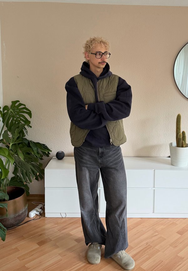 Personne aux cheveux blonds bouclés, portant des lunettes et une moustache, debout les bras croisés, vêtue d'un gilet vert, d'un sweat à capuche noir, de jeans gris larges et de chaussons à l'intérieur.