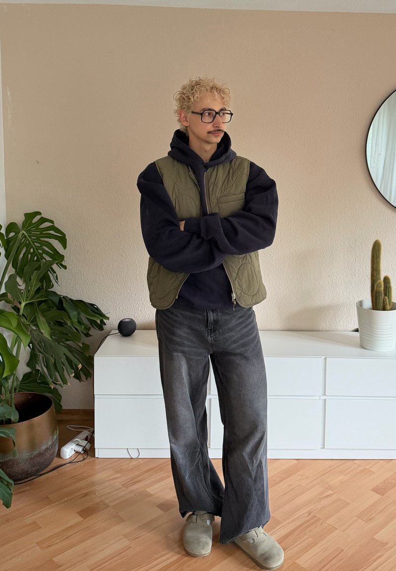 Personne aux cheveux blonds bouclés, portant des lunettes et une moustache, debout les bras croisés, vêtue d'un gilet vert, d'un sweat à capuche noir, de jeans gris larges et de chaussons à l'intérieur.
