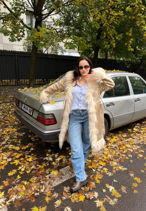 Une femme portant des lunettes de soleil et un manteau bordé de fourrure pose à côté d’une voiture argentée, dans une rue couverte de feuilles en automne.