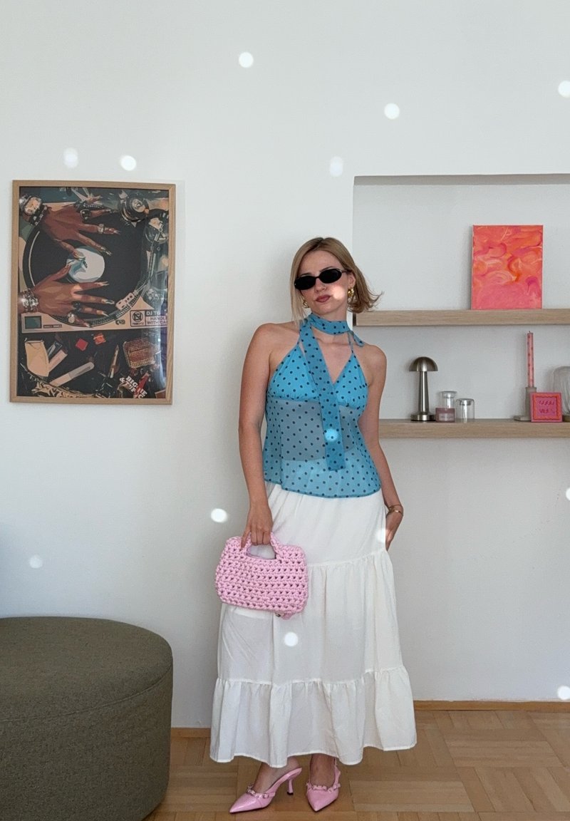 Femme en haut à pois bleu à col halter, jupe blanche à volants, talons roses et sac à main rose au crochet posant à l'intérieur avec une œuvre d'art murale et des étagères derrière.