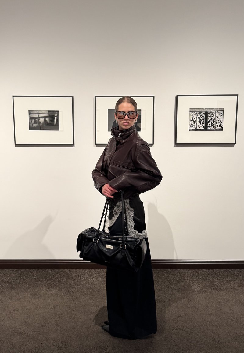 Ženska v temni usnjeni jakni in velikih sončnih očalih drži črno torbico in stoji pred tremi okvirjenimi črno-belimi fotografijami.