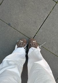 Brune Puma-sneakers med hvite detaljer og sømmer, kombinert med løse hvite bukser, står på en teksturert grå asfalt.