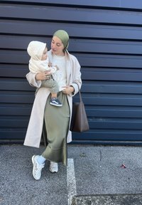 Γυναίκα φορώντας ένα μπεζ παλτό και ελιόχρωμη φούστα κρατά ένα παιδί ντυμένο με μια κρέμα κουκούλα. Μια καφέ τσάντα και λευκά αθλητικά ολοκληρώνουν το σύνολο μπροστά σε μπλε φόντο.