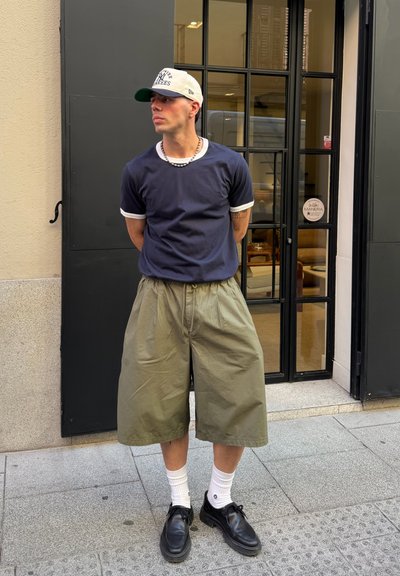 Joven con camiseta azul marino, pantalones cortos beige anchos, calcetines blancos, zapatos negros y gorra de béisbol blanca, de pie en la acera cerca de una puerta de cristal.