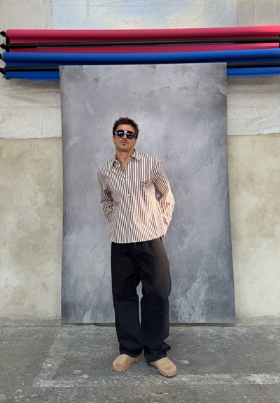 Camisa de botones a rayas en blanco y marrón, pantalones holgados negros y zuecos beige, con gafas de sol oscuras contra un fondo gris texturizado.