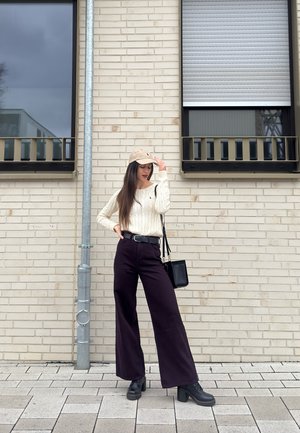 Mujer con gorra beige, suéter crema, pantalones anchos negros y botas, de pie sobre una acera pavimentada frente a una pared de ladrillo clara.
