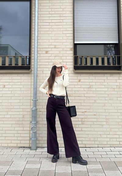 Mujer con gorra beige, suéter crema, pantalones anchos negros y botas, de pie sobre una acera pavimentada frente a una pared de ladrillo clara.