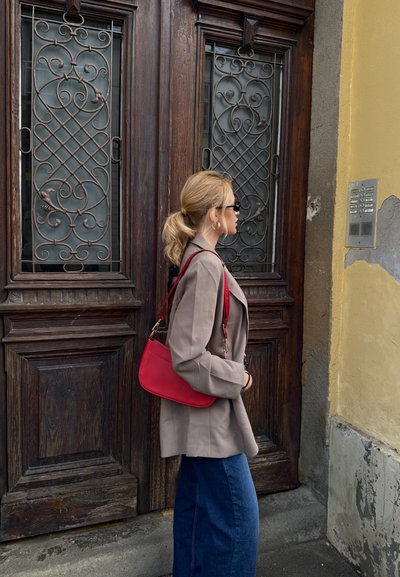 Mujer con coleta rubia, blazer beige, bolso rojo y gafas de sol, de pie junto a grandes puertas de madera con rejas metálicas ornamentadas.