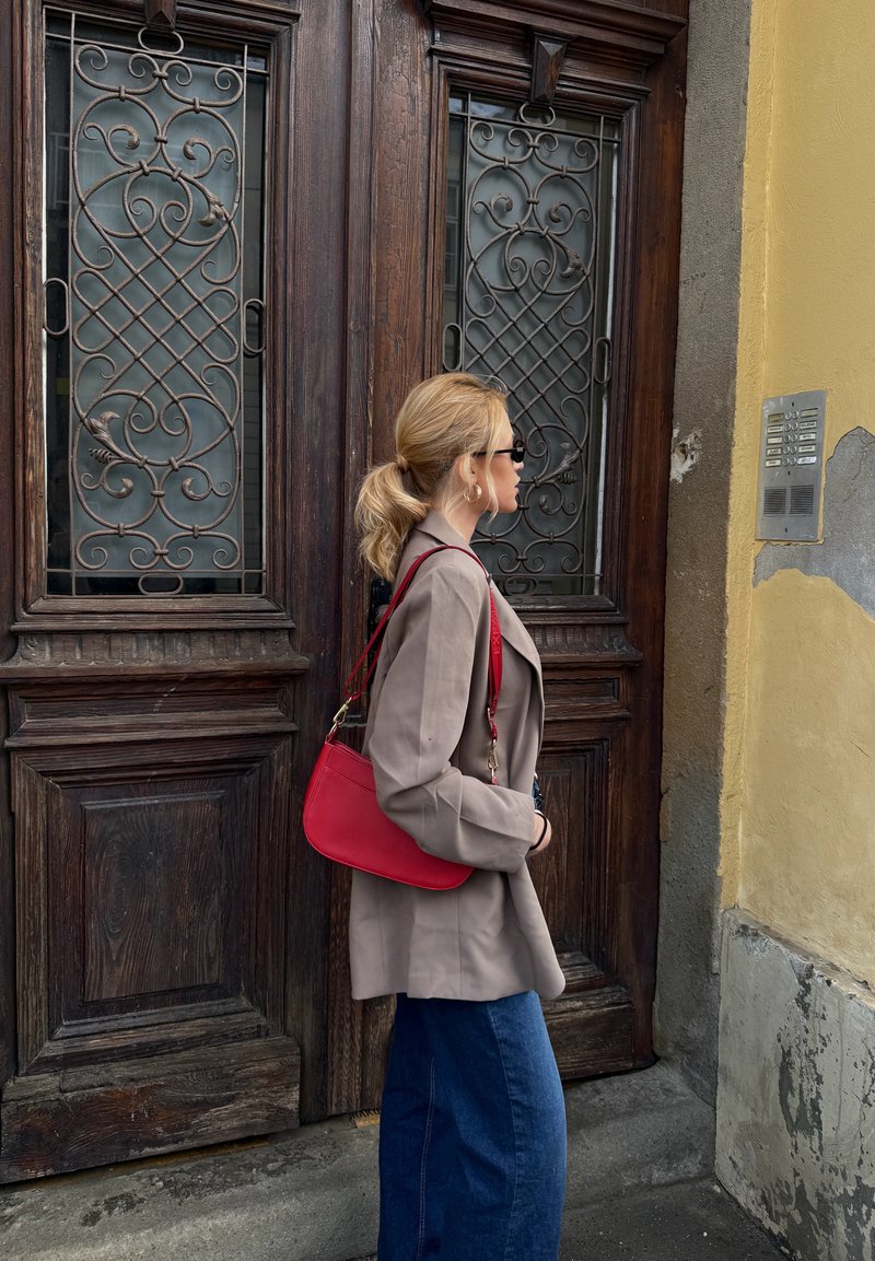 Woman with blonde ponytail, beige blazer, red purse, and sunglasses standing beside large wooden doors with ornate metal grilles.
