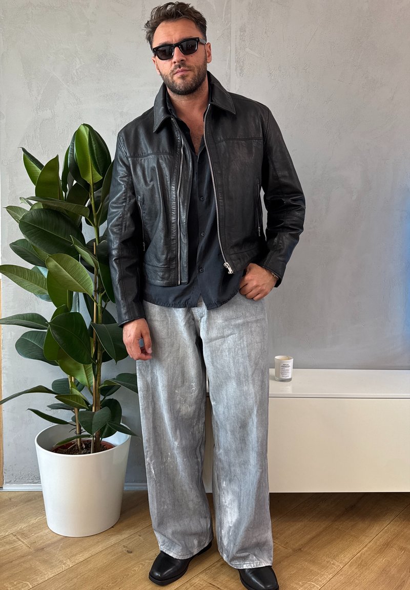 Veste bomber en cuir noir portée sur une chemise sombre, associée à un pantalon large gris clair. Lunettes de soleil foncées et chaussures noires. Cadre intérieur avec plante.