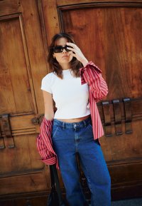 T-shirt bianca aderente, camicia oversize rossa e bianca a righe, jeans in denim blu a vita alta e occhiali da sole neri. Fondo in legno.