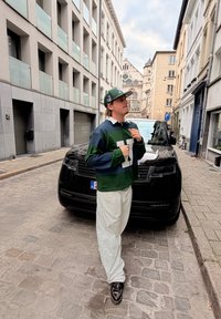Osoba s prúžkovaným polo tričkom v zelenej a námorníckej modrej farbe s bielym golierom, bielymi nohavicami a čiernymi topánkami, stojaca vedľa čierneho auta na dlažobnej ulici.