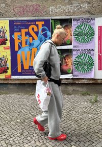 Grå træningsdragt med løs pasform, røde sneakers og en sort håndtaske. Bærer en hvid taske med lyserødt indre, som indeholder en plante. Plakater i baggrunden.